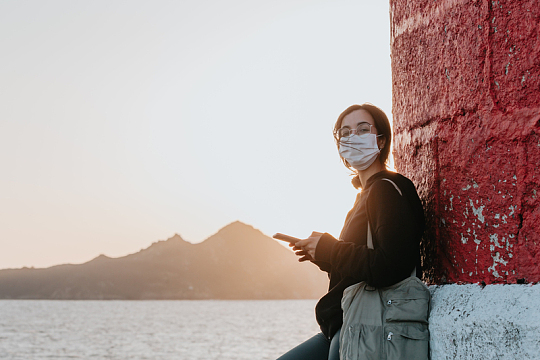 夕阳下海边靠在墙上的黑衣戴口罩女性